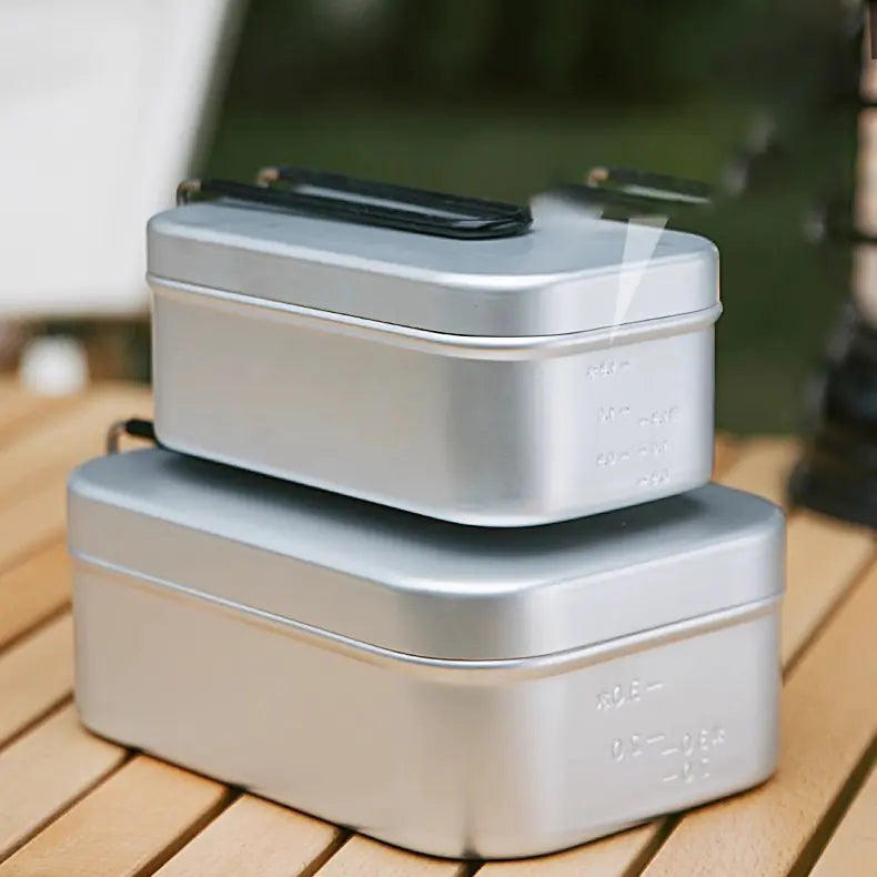 Two metallic german cooking boxes stacked on a wooden surface with a blurred outdoor background